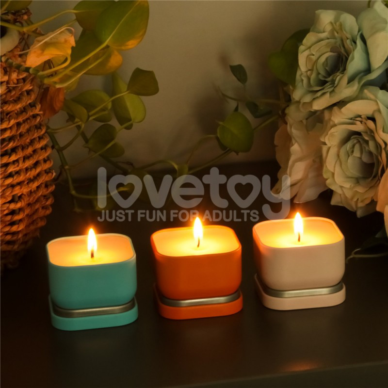 Набір масажних свічок Hush у блакитній, помаранчевій і світло-рожевій банках, запалені, на фоні квітів